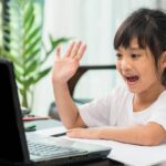little girl smiling and rasing her hand in front of laptop